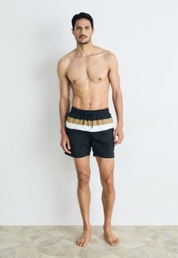 Schwarze Badehosen mit einem weißen und goldenen horizontalen Streifen, ausgestattet mit einem elastischen Bund und Zugband zur Anpassung.