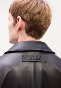 Black leather jacket featuring a wide collar; embossed "HUGO" logo with square accents on the back. Smooth texture and fitted design.