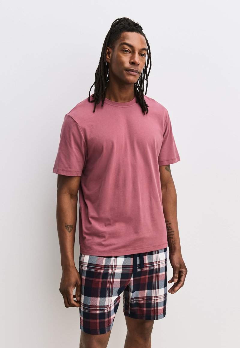 Homme avec des dreadlocks portant un t-shirt rose à manches courtes et un short à carreaux bleu marine, blanc et rouge, debout devant un fond blanc uni.