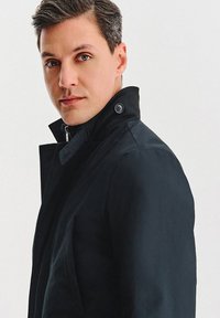 Czarny płaszcz trencz z wysokim kołnierzem, wyposażony w zamek błyskawiczny z przodu i ozdobny guzik. Materiał wydaje się gładki i matowy, podkreślając jego dopasowany wygląd.