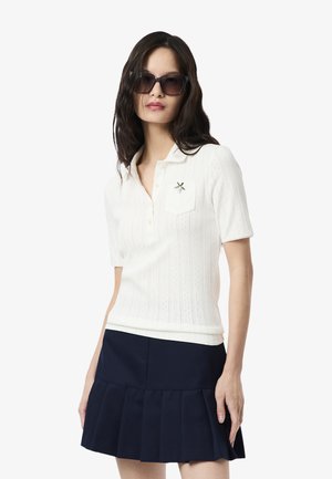 Polo shirt blanc à manches courtes avec un design texturé et une libellule brodée sur la poche, assorti d'une jupe plissée bleu marine.