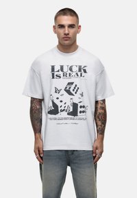 Witte katoenen t-shirt met een gedurfde zwarte grafiek met de tekst "LUCK IS REAL," een patroon van dobbelstenen en vlinders, korte mouwen, losse pasvorm.