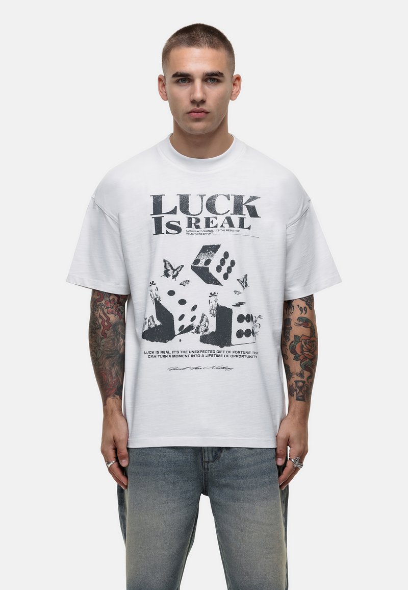 Witte katoenen t-shirt met een gedurfde zwarte grafiek met de tekst "LUCK IS REAL," een patroon van dobbelstenen en vlinders, korte mouwen, losse pasvorm.