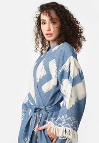 Modrá žakárová župan s abstraktnými bielymi vzormi, textúrovaná látka, široké rukávy, opasok v páse a strapcové okraje.