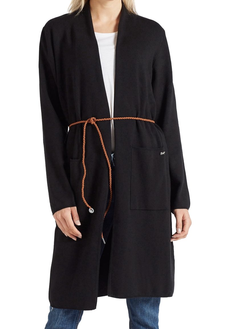 Femme portant un long cardigan noir avec des poches devant, ceinturé à la taille d'une fine ceinture brune tressée, sur une chemise blanche et un jean bleu.