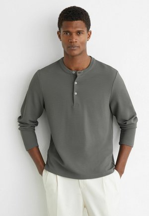 Młody mężczyzna ubrany w szarą, długorękawową koszulę henley i białe spodnie, stojący z rękami w kieszeniach na tle białej ściany.