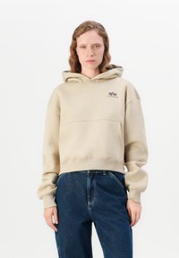 Felpa cropped beige con grande tasca frontale, caratterizzata da un piccolo logo marrone sul petto e polsini aderenti. Abbinata a jeans blu scuro.