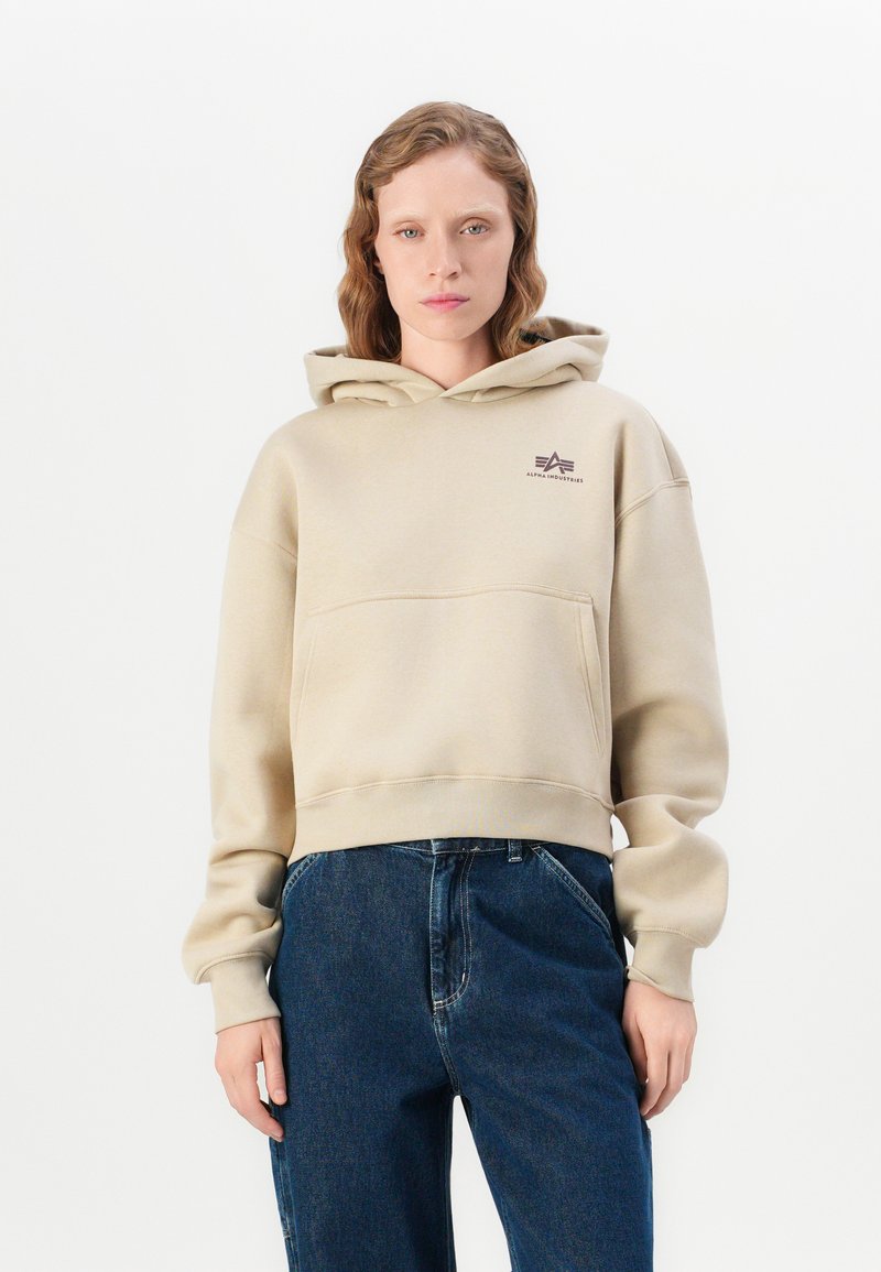 Felpa cropped beige con grande tasca frontale, caratterizzata da un piccolo logo marrone sul petto e polsini aderenti. Abbinata a jeans blu scuro.
