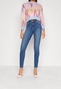 Suéter tejido colorido con patrones geométricos en rosa, azul y beige, combinado con jeans ajustados azules y tacones rosa claro.