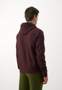 Haut à capuche bordeaux avec une coupe décontractée, capuche ajustable et ourlet côtelé, associé à un pantalon vert. Texture lisse, manches longues et sans motifs visibles.