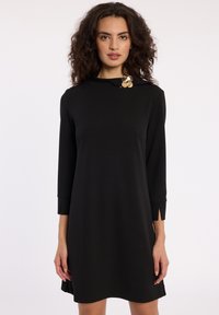 Robe noire mi-longue avec des manches trois-quarts, dotée d'un col montant et de poignets fendus. Un accent circulaire doré au niveau du col ajoute du détail.