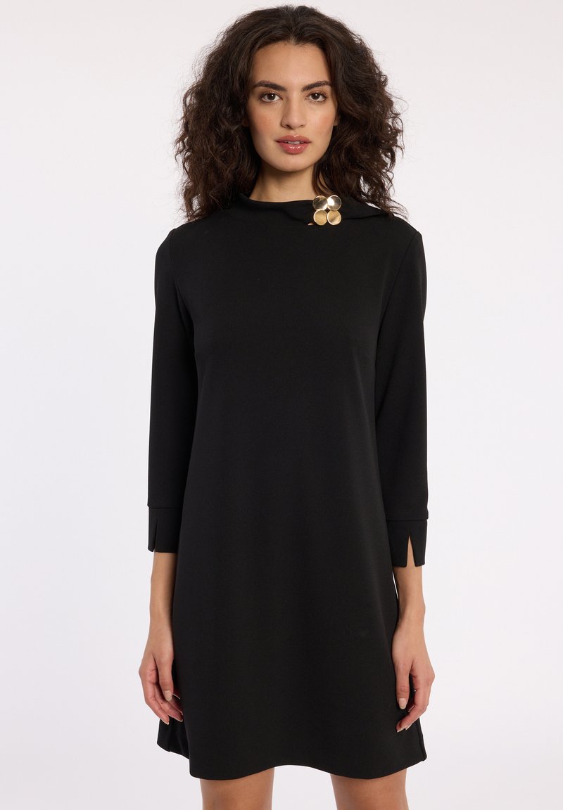 Robe noire mi-longue avec des manches trois-quarts, dotée d'un col montant et de poignets fendus. Un accent circulaire doré au niveau du col ajoute du détail.