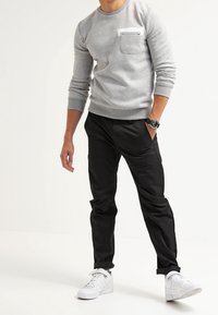 Felpa grigia a girocollo con tasca bianca, abbinata a pantaloni neri a gamba stretta e sneakers bianche. Tessuto morbido e vestibilità casual.