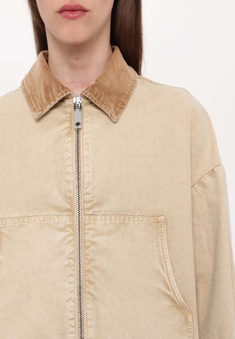 Beige jas met een zacht corduroy kraag, ritssluiting aan de voorkant en grote borstzak. De stof lijkt glad met een subtiele glans.