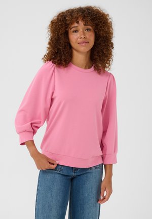 Vrouw met krullend haar die een roze blouse met lange mouwen en een blauwe spijkerbroek draagt, staand tegen een effen lichte achtergrond.