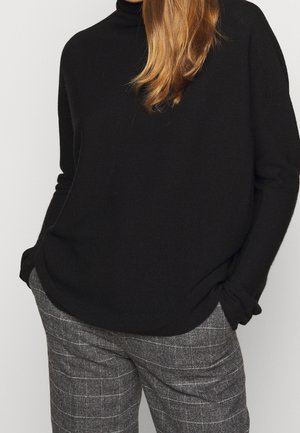 Person iført sort rullekrave-pullover med hænderne i lommerne og grå ternede bukser mod en ensfarvet baggrund.