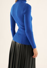 Pull à col roulé bleu côtelé avec une silhouette ajustée et des manches longues, associé à une jupe plissée noire fluide.