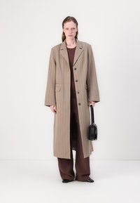 Long beige striped coat with single-breasted buttons and front pockets, worn over a brown outfit, holding a small black handbag.