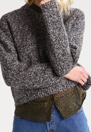 Personne portant un pull en tricot noir et blanc sur une chemise verte à motifs et un jean bleu, bras plié avec la main près du visage.