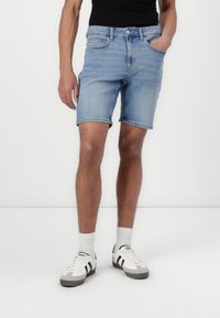 Șorturi din denim albastru deschis, cu margini răsucite, două buzunare frontale și închidere cu nasture, purtate cu adidași albi și șosete rayate.