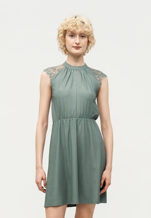 Kvinde med kort krøllet blondt hår iført en ærmeløs salviegrøn kjole med blondekapsler, stående foran en enkel lys baggrund.