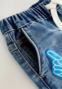 Jeans in denim blu con vita elastica, tasca frontale e una toppa a forma di mano in schiuma blu che punta verso l'alto sulla gamba.