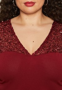 Vestido burdeos con un escote en V profundo y un intrincado encaje con lentejuelas en los hombros, presentado en un tejido suave y ligeramente texturizado en la parte inferior.
