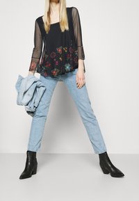 Sort blomsterbroderet bluse med gennemsigtige lange ærmer, parret med lyseblå jeans og sorte ankelstøvler, der viser en afslappet pasform og lagdelt design.