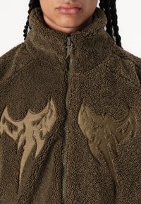Brun fleecjacka med hög krage, med två stora beige broderade mönster på framsidan. Dragkedjestängning och texturerad tyg.