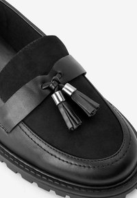 Schwarze Lederloafer mit einer Vorderseite aus Wildleder, einem breiten Riemen mit Quasten und metallischen Akzenten an den Quastenbasen. Strukturierte Gummisohle.