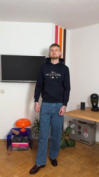 Μπλε φούτερ με κουκούλα με το λογότυπο "Hilfiger", μπλε παντελόνια με φαρδιά πόδια με σχέδιο, καφέ παπούτσια και μοντέρνο εσωτερικό χώρο με πολύχρωμη διακόσμηση και φυτά.