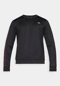 Sudadera negra de manga larga con franjas laterales rojas con patrón y un logo bordado en verde en el pecho. Fabricada en tejido ligero.