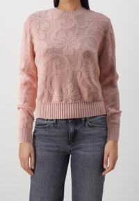 Pull en maille rose présentant un motif floral en relief, avec des poignets et un ourlet côtelés. Porté avec un jean gris, affichant une coupe et un style décontractés.