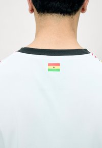 Arrière du cou d'une personne portant une chemise blanche avec un petit drapeau du Ghana imprimé sous le col noir.