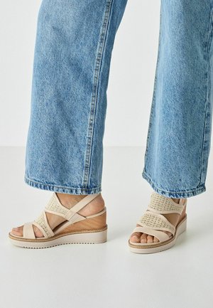 Beżowe sandały na klinie z krzyżującymi się paskami z plecionymi akcentami, gładkim drewnianym obcasem i teksturowaną podeszwą, zestawione z jasnoniebieskimi szerokimi spodniami.