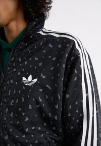 Black zip-up jacket with a leopard print pattern, featuring white stripes along the shoulders and a small white Adidas logo on the chest.