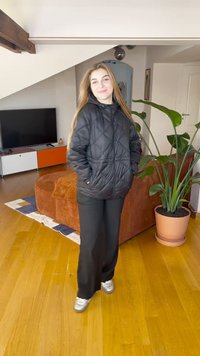 Black quilted jacket with a hood, worn over black pants and silver sneakers. Indoor setting with a plant and sofa in the background.