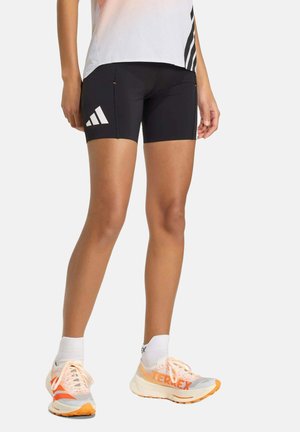 Mujer con pantalones cortos deportivos negros de Adidas, calcetines blancos y zapatillas de correr blancas y naranjas, de pie sobre un fondo blanco.