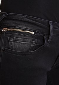 Jeans de mezclilla negra con un bolsillo con cremallera, costuras sutiles y un diseño ajustado. La tela tiene una textura suave y un brillo mínimo.