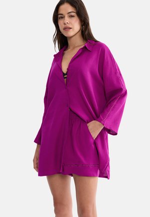 Kvinde iført løs, magentarød skjorte og matchende shorts med lommer, stylet over en sort top, stående foran en ensfarvet baggrund.