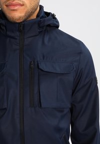 Marinblå huva jacka med flera framfickor, slät textur och frontdragkedja. Har justerbar huva för passform.