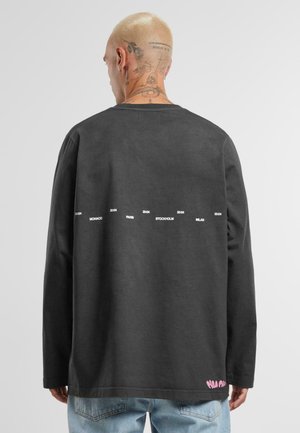 T-shirt à manches longues gris foncé avec un col rond. Le dos présente un texte blanc faisant référence à des villes et à des horaires ; logo rose en accent sur l'ourlet.