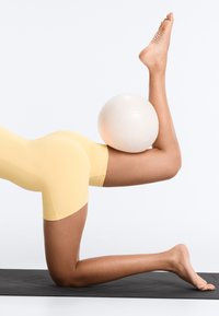 Personne en short jaune agenouillée sur un tapis noir, tenant un ballon d’exercice blanc entre la cuisse et le mollet plié, un pied pointé vers le haut.