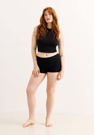 Junge Frau mit langen roten Haaren steht barfuß in schwarzem ärmellosem Oberteil und Shorts vor weißem Hintergrund und zeigt Tätowierungen auf den Armen.