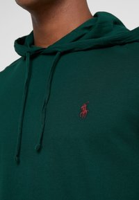 Grön bomullshuvtröja med en framficka och dragskor. Har en liten vinröd broderad logotyp på vänster bröst.