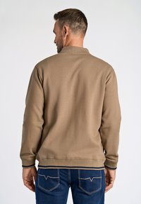 Felpa beige con collo alto, polsini e orlo a coste, indossata sopra jeans blu scuro. Tessuto liscio, design minimale, senza pattern visibili.