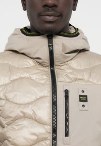 Beige gewatteerde jas met een capuchon, voorzien van een rits aan de voorkant, zwarte accenten en een logolabel op de borstzak. Soepele, glanzende textuur.