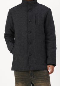 Tmavě šedý kabát z vlněné směsi s vysokým límcem, zapínáním na knoflíky a bočními kapsami. Vzor rybí kosti s detailem jedné kapsy na hrudi.