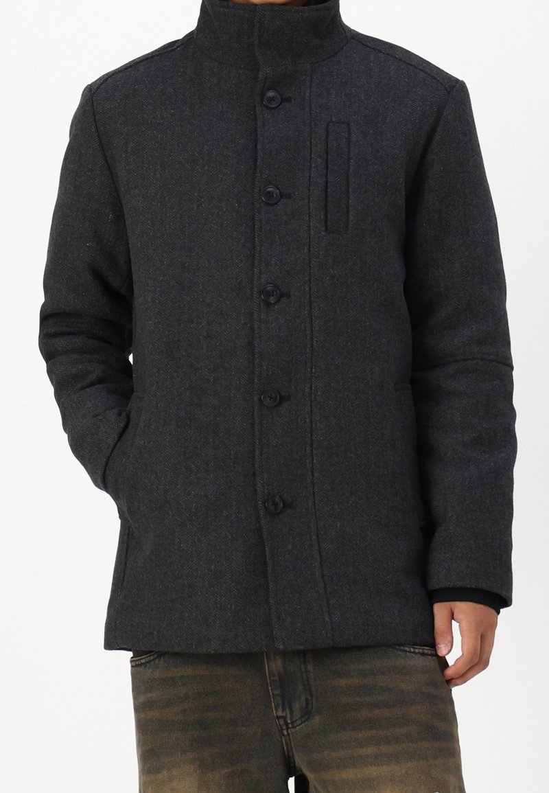 Tmavě šedý kabát z vlněné směsi s vysokým límcem, zapínáním na knoflíky a bočními kapsami. Vzor rybí kosti s detailem jedné kapsy na hrudi.
