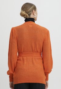 Oranžinė nėriniuota kardiganas su diržo juosta, ilgomis rankovėmis ir laisva siluetu. Tekstūruota medžiaga su minkštu ir šiek tiek pūkuotu išvaizda.